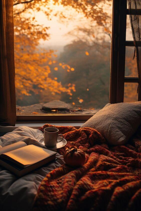 Lit devant une fenêtre avec un livre et une tasse de thé devant un paysage d'automne en fin de journée